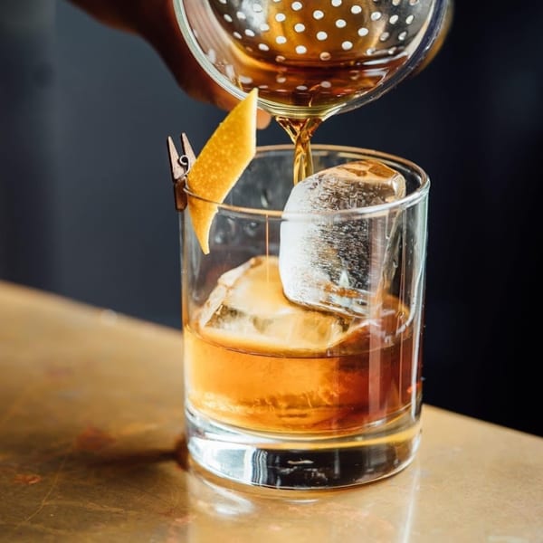 Bartender pouring a dark amber cocktail from a strainer into a glass with ice and an orange peel.