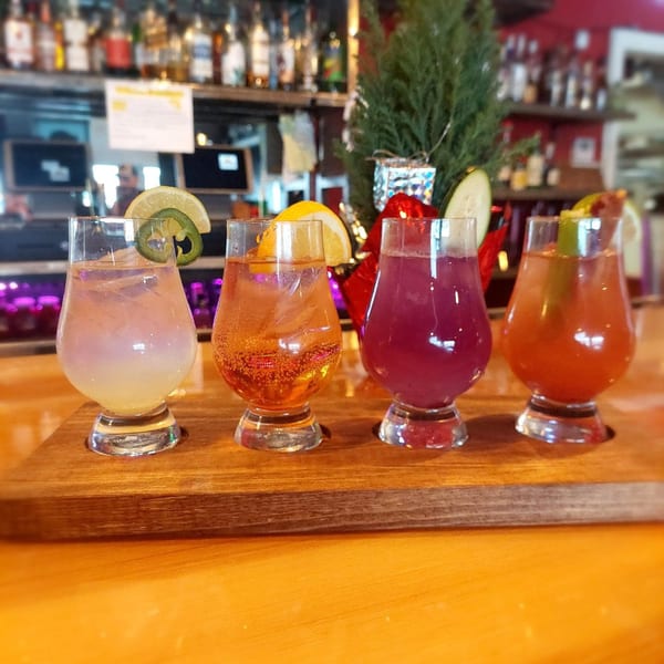 Four colorful cocktails, garnished with citrus, cucumber, and jalapeño, on a wooden tray at F325 BBQ.