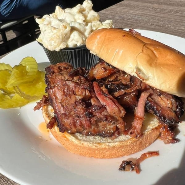 F325 BBQ brisket sandwich with a side of macaroni salad and pickles on a white plate.
