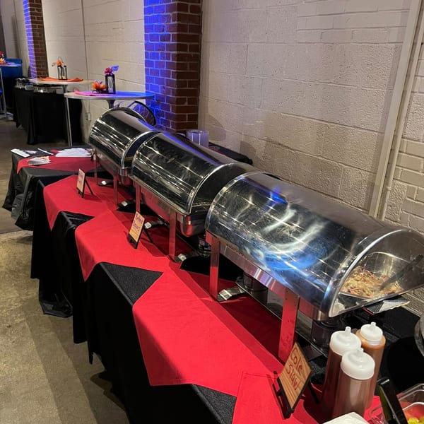 A BBQ buffet line with three chafing dishes, red and black tablecloths, and sauce bottles.