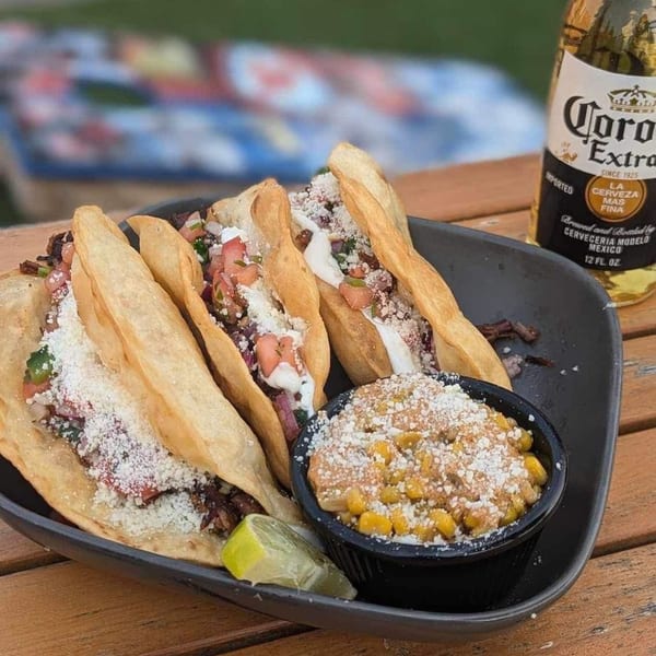 Three F325 BBQ tacos with elote and a Corona Extra beer on a wooden table.