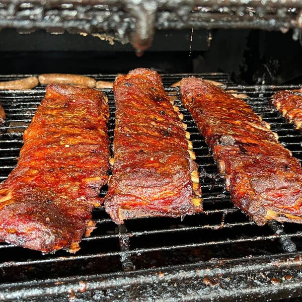 Three racks of F325 BBQ ribs and sausages cooking on a dark, smoky grill.